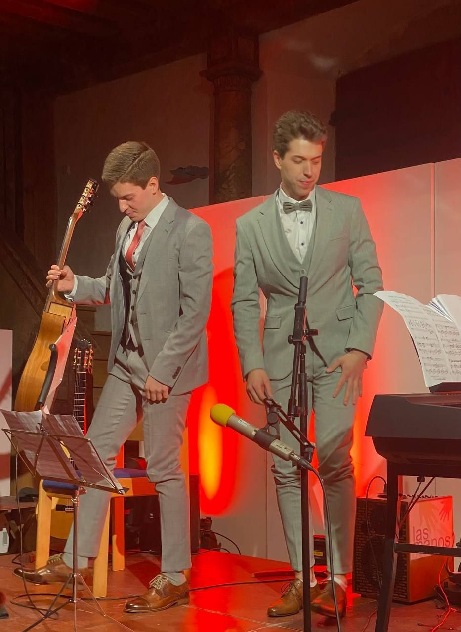 Two men in grey suits standing on a stage with musical instruments and microphones.