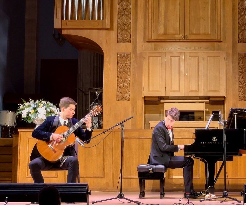 Two musicians performing on stage, one playing classical guitar and the other on a grand piano.